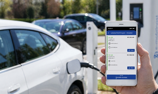 Ruka drži pametni telefon s prikazom aplikacije za polnjenje električnega vozila, medtem ko se v ozadju električni avtomobil polni na polnilni postaji